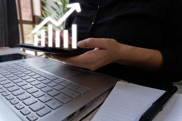 Businesswoman in front of a computer holding a cell phone and above it there is a virtual graph with an arrow going up
