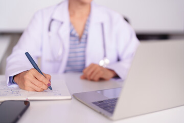 Experienced female doctor writes patient notes medical records consultation details on office desk with laptop smartphone healthcare provider at clinic