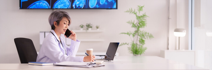 Asian doctor physician woman professional consults patients remotely via phone writes medical notes laptop computer modern office clinic setting healthcare technology