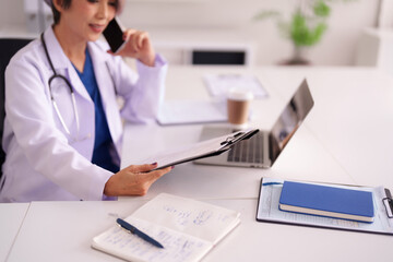 Adult Asian Female Physician Manages Healthcare Administration Duties From Modern Clinic Office Space Utilizing Phone Laptop Tools