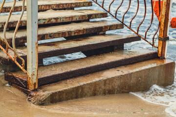 Rusty metal steps transition from a worn landing to the sandy beach, with gentle ocean waves lapping against the base, creating a tranquil coastal atmosphere.