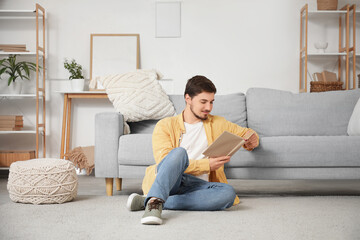 Fototapeta premium Young man reading book on carpet near sofa at home