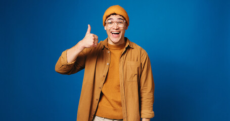 Naklejka premium Happy man, portrait and fashion with thumbs up in studio for good choice on a blue background. Male person, model or gen z with smile, like emoji or yes sign for style or clothing on mockup space