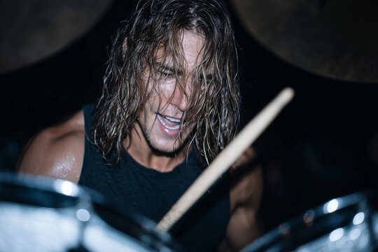 Man with long wet hair drumming passionately in black tank top, intense live performance under dramatic lighting