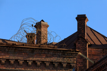 Weathered red-brick wall of the Kresty prison in Saint Petersburg with chimneys, rusted barbed wire, and aged metal roof under clear sky.