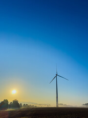 Obraz premium A Serene and Breathtaking Sunrise Over a Wind Turbine, Lovingly Surrounded by Misty, Lush Fields that Stretch Out into the Horizon, Embodying the Beauty of Nature and Sustainable Energy