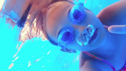 Montage of a young girl swimming underwater, wearing blue goggles and a colorful swimsuit, playing and making bubbles in a brightly lit pool