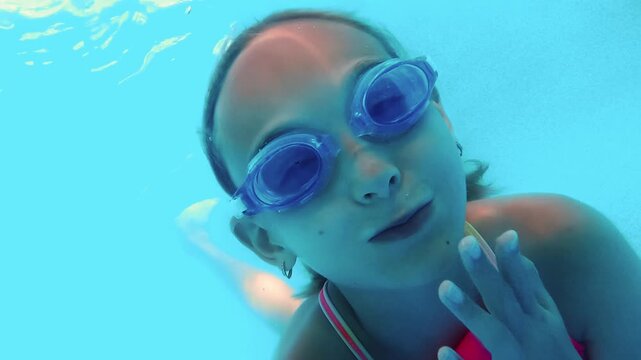 Young girl wearing swimming goggles playing underwater, creating funny faces and blowing bubbles with playful joy and carefree expression in bright blue pool water