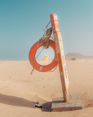 An emergency lifebuoy ring with an SOS sign stands isolated in a wide, sandy beach or desert landscape under a clear blue sky. The minimalistic scene emphasizes solitude, safety, and the stark contras