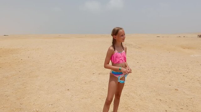 Young girl wearing pink bikini standing alone on vast sandy landscape, clutching swimming goggles while surrounded by expansive desert terrain under bright cloudless sky
