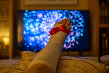 Broken foot with a red bow rests on a pillow in front of a television displaying vibrant fireworks during the christmas holiday season, suggesting themes of resilience and recovery