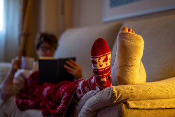 Woman enjoys a quiet christmas evening reading a book and drinking a hot beverage while recovering from a broken foot, wearing a festive christmas sock and wrapped in a warm blanket