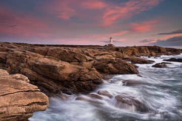 Lorient Morbihan Waves rushing against the rocks with a lighthouse in the background during sunset.