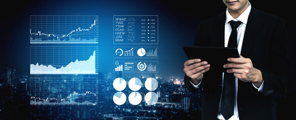 A businessman in a suit focuses on analyzing financial data trends using a digital tablet, surrounded by stock market charts against a lit cityscape. Impute
