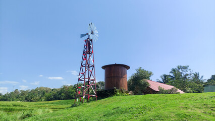 Obraz premium High-quality photo of a rural landscape with windmill, water tank, and house in green rice fields under blue sky. Perfect for agriculture, renewable energy, countryside, and environmental projects.