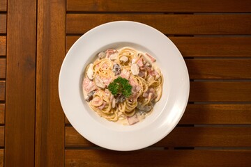 Creamy Ham and Mushroom Pasta. Classic Italian spaghetti with a rich, white sauce, diced ham, and sliced mushrooms, garnished with parsley. Comfort food on a wooden table.