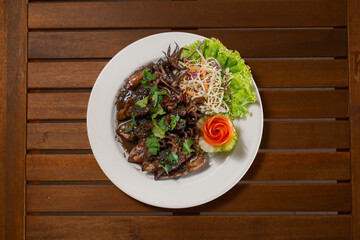 Thai Stir-Fried Squid in Black Ink Sauce (Muek Pad Nam Dam). A unique and savory seafood delicacy from Thailand served on a plate with fresh greens on a wooden table. Gourmet Asian cuisine.