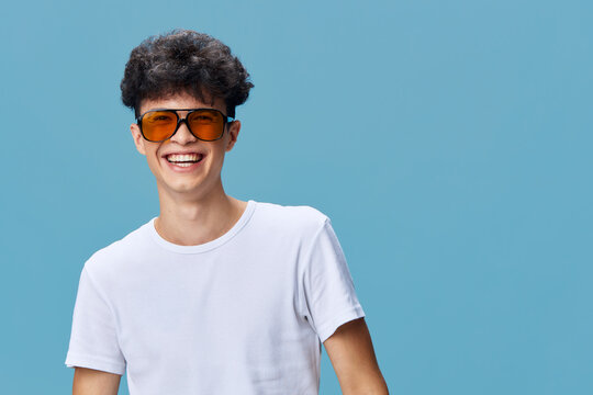 smiling man wearing orange sunglasses and white t-shirt, joyful young adult with curly hair against blue background expressing happiness and style in casual attire