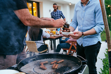 A group of friends and family barbecue together in the evening on the terrace in front of a large modern house. 