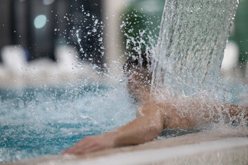 Man standing under a hydrotherapy waterfall in a spa pool, receiving water massage on his head and shoulders.