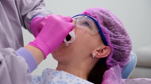 Dentist wearing pink gloves and protective glasses placing an optragate lip retractor in patient's mouth, preparing for a teeth whitening or other dental procedure