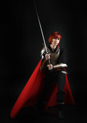 Fantasy warrior woman model in historical medieval armour costume, red fairytale cloak, cinematic portrait of strong fantasy knight holding sword. Epic standing pose, dark studio background