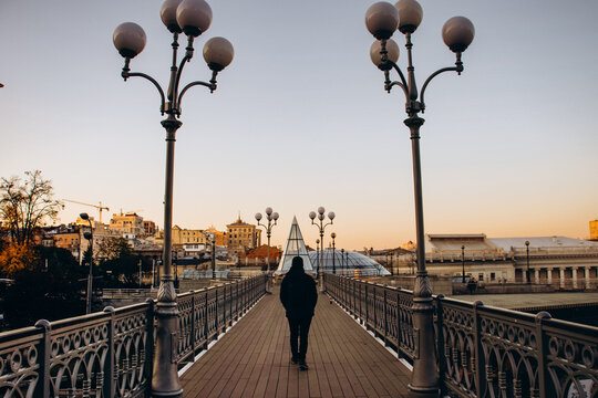 A lone figure walks away from the camera on a decorative pedestrian bridge in Kyiv at sunset, with ornate street lamps lining the path and a panoramic view of the city skyline in the warm light