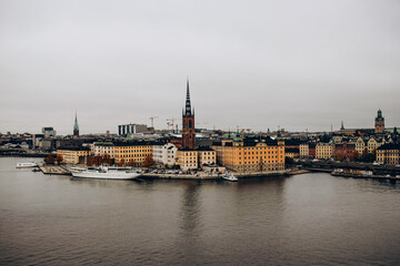 A panoramic view of the Stockholm cityscape, showcasing the historic buildings of the old town, Gamla Stan, against the backdrop of a gray sky with a cruise ship in the harbor.