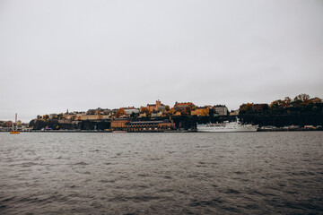The hilly waterfront of Stockholm's Södermalm district, with historic buildings and a large cruise ship docked along the shore on a cloudy, autumnal day.