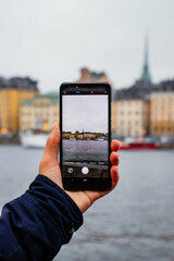 A person holds a smartphone, taking a photo of the Stockholm skyline across the water. A representation of modern travel and city exploration.