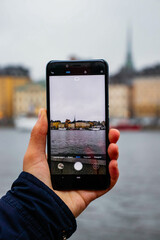 A person holds a smartphone, taking a photo of the Stockholm skyline across the water. A representation of modern travel and city exploration.