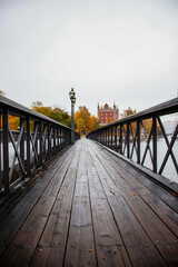 Obraz premium A wooden bridge leading across a river to a historic red building on the opposite bank, surrounded by colorful autumn trees in Stockholm, Sweden.
