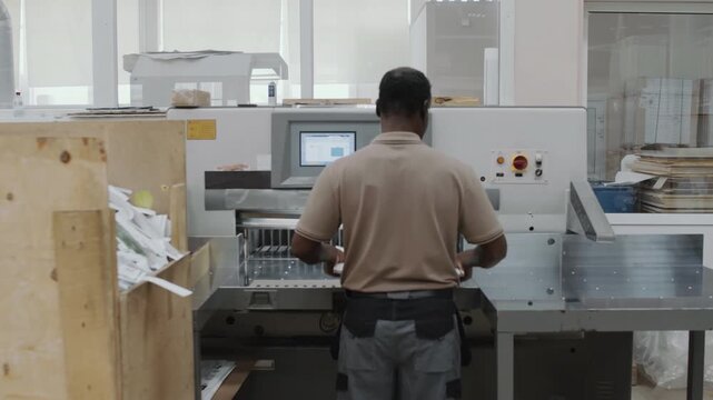 Slow motion dolly in rear view of mature Black man using paper cutter in modern printing house