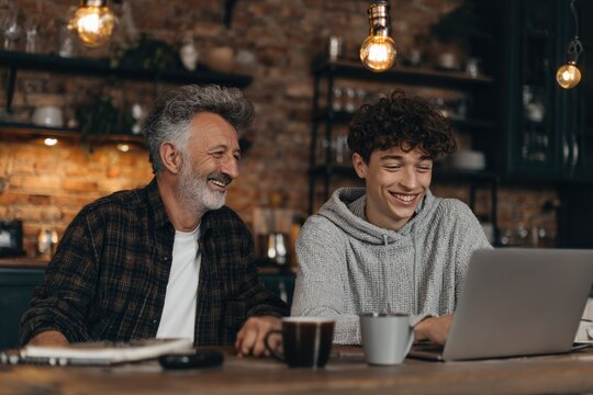 elderly man and his teenage grandson sitting at a wooden table in a countryside home, editing a video on a laptop together, laughing and bonding
