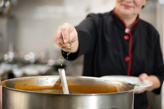 Close up of a chef in black uniform adding fresh herbs to a large pot of soup in a professional kitchen. Concept of gastronomy, seasoning, culinary preparation, and hospitality industry