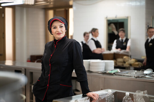 Portrait of a female chef in a black uniform with red details smiling in a professional restaurant kitchen. Image highlights hospitality staff, confidence, and culinary professionalism.