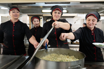 Group of chefs in black uniforms preparing rice in a large pot in a commercial kitchen. Female cooks stirring and adding herbs while working as a team. Concept of teamwork, gastronomy, and hospitality
