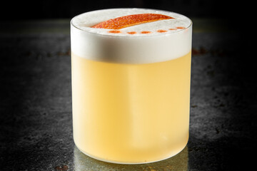 Elegant cocktail with frothy foam, orange slice garnish, and bitters drops, served in a clear glass on a dark textured surface.