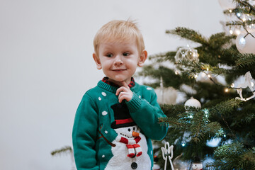 Little boy with finger on chin expressing thoughtful happy emotion at Christmas tree