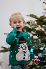 Little boy in green Christmas shirt with snowman by Christmas tree expressing surprise emotion