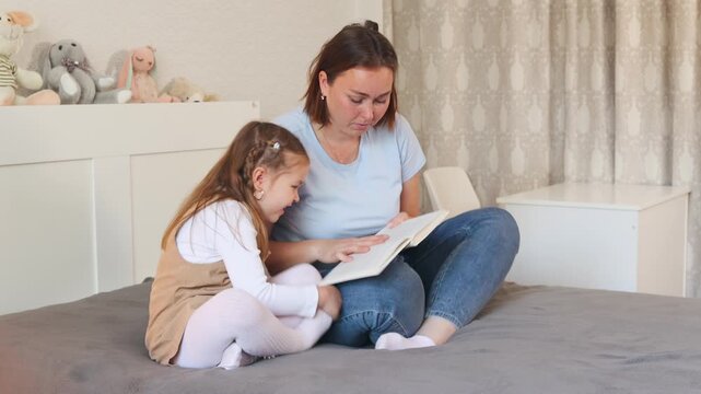 Little Caucasian girl learns to read with her mother while sitting on a bed in a bright room. Early education and preschool development at home. Wide shot.