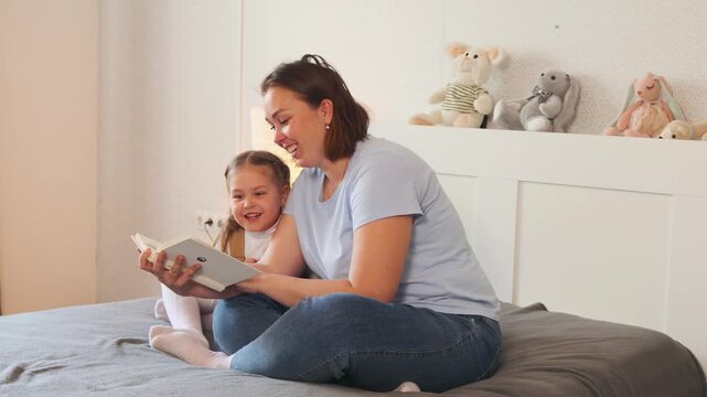 Young child follows along while sitting with her mother reading books. Early childhood literacy and emotional support. Wide shot.