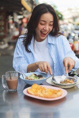 Smiling Asian woman eating Thai breakfast at local street food market, authentic lifestyle and happiness concept.