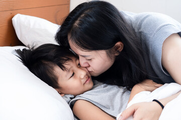 Asian mother spending quality time with her little boy, playing and kissing him while lying on a white bed, enjoying love, bonding, relaxation, and happy family moments together in the bedroom