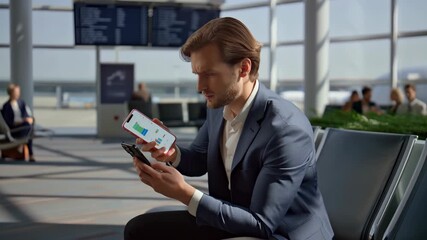 Businessman sitting in airport terminal using two smartphones, checking graphs and data while waiting for his flight, with other passengers and flight information display in background - Powered by Adobe
