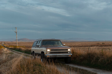 Fototapeta premium Vintage SUV with illuminated headlights in Irish countryside grassland at dusk