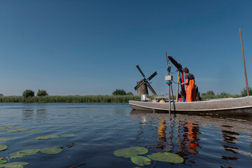 Research boat with drilling equipment on Dutch waterway with historic windmill and lily pads visible in background
