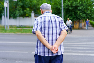 Elderly man waits to cross a busy street with hands clasped behind his back during daylight