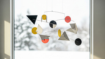 Geometric mobile art suspended against a winter landscape backdrop, showcasing an artistic display. The mobile features an array of shapes, including triangles, circles, and abstract forms.
