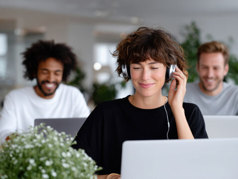 Woman enjoying music while working on laptop in office with coworkers